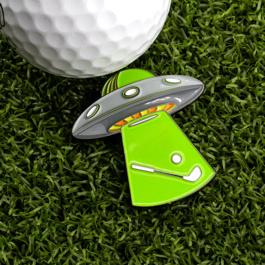The Albatross Abduction Ball Marker by Ballmarkers.com rests on artificial grass, its UFO shape and golf club and ball beam design celebrating those elusive albatross shots on the course.