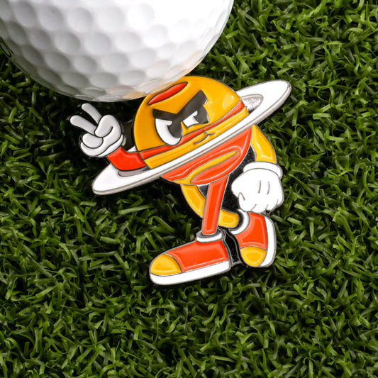 A cartoon character looking like a yellow planet flashes a peace sign on green turf beside a white golf ball, sporting red shoes and gloves—set for a "Mission to Pars" with the Saturn Out Ball Marker by Ballmarkers.com.