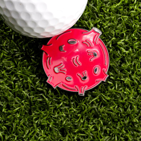 The Terra Rossa Ball Marker by Ballmarkers.com—a red, round marker with a virus-inspired design—sits on artificial green grass beside a white golf ball.