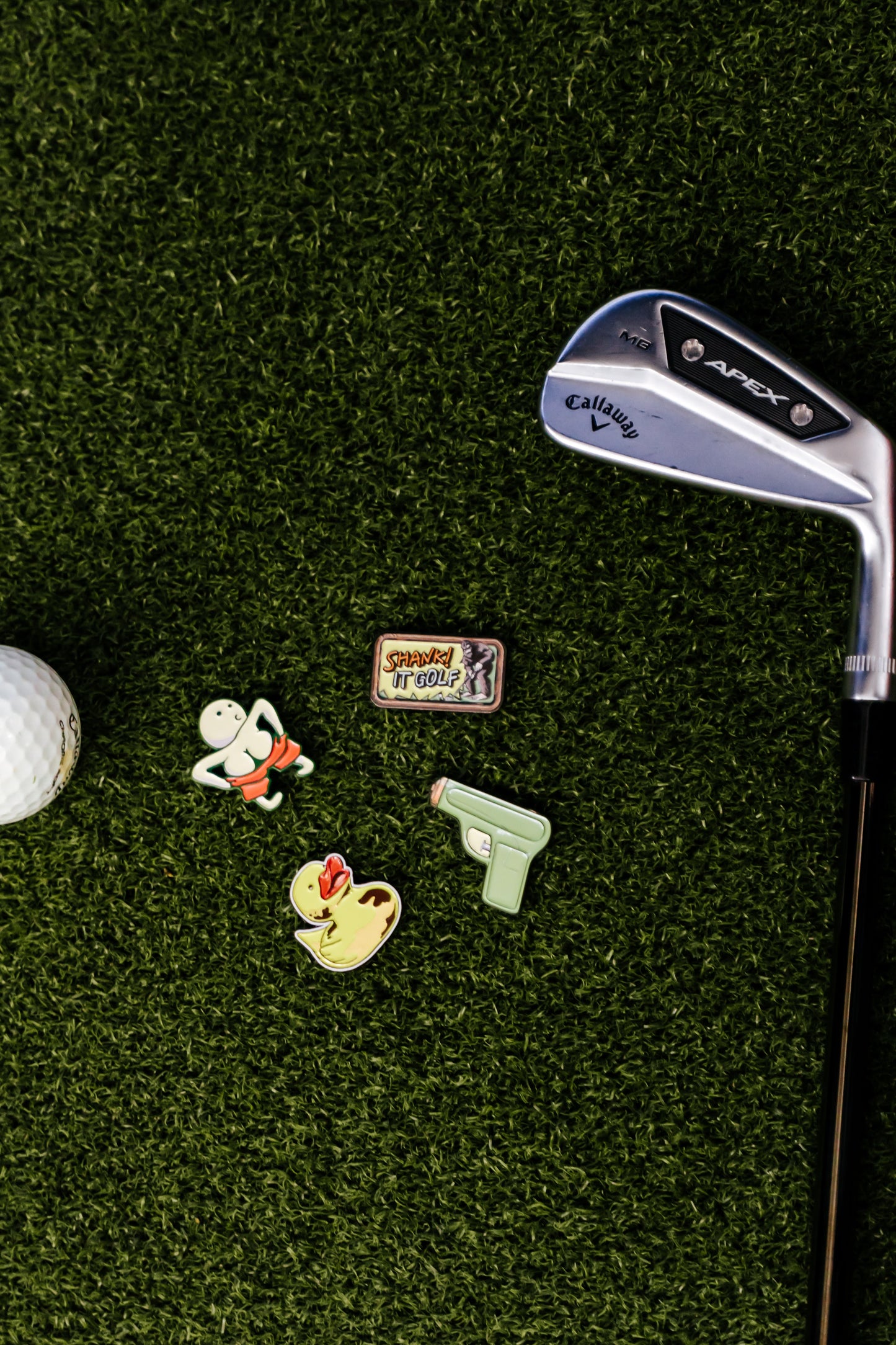 A Callaway golf club, a premium metal golf ball, and four Shank it Golf ball markers—including the Ducky Ball Marker—are displayed on green artificial turf.