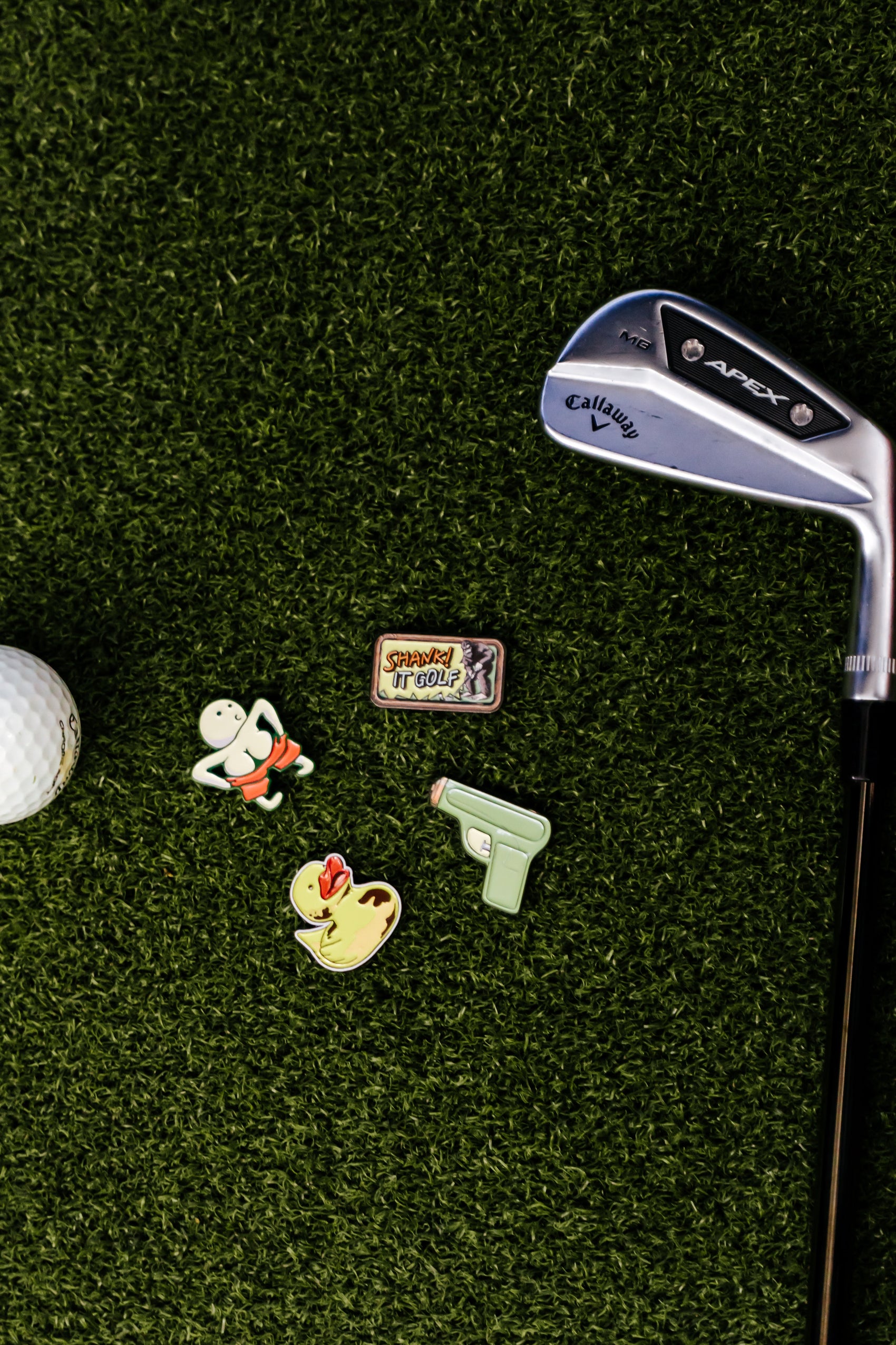 A Callaway golf club, a premium metal golf ball, and four Shank it Golf ball markers—including the Ducky Ball Marker—are displayed on green artificial turf.