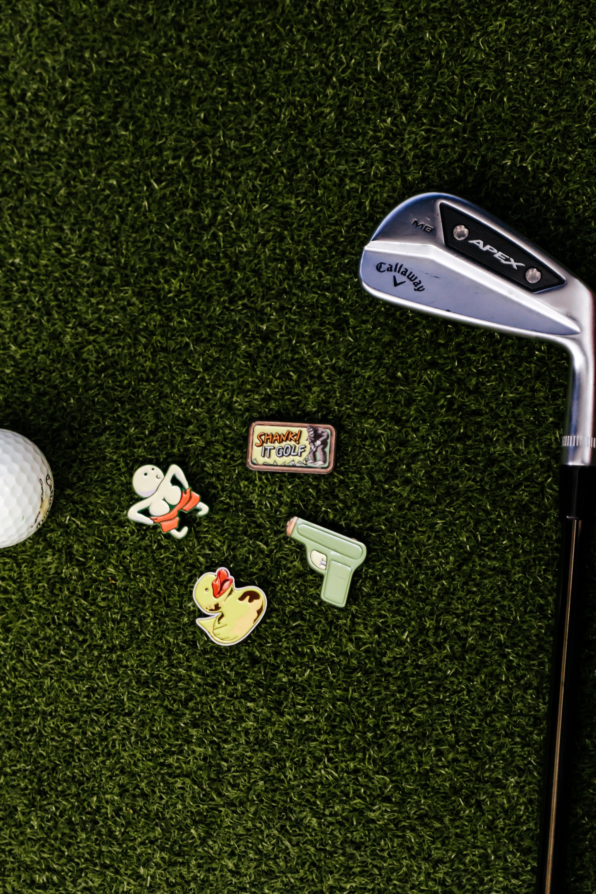 A Callaway golf club, a premium metal golf ball, and four Shank it Golf ball markers—including the Ducky Ball Marker—are displayed on green artificial turf.