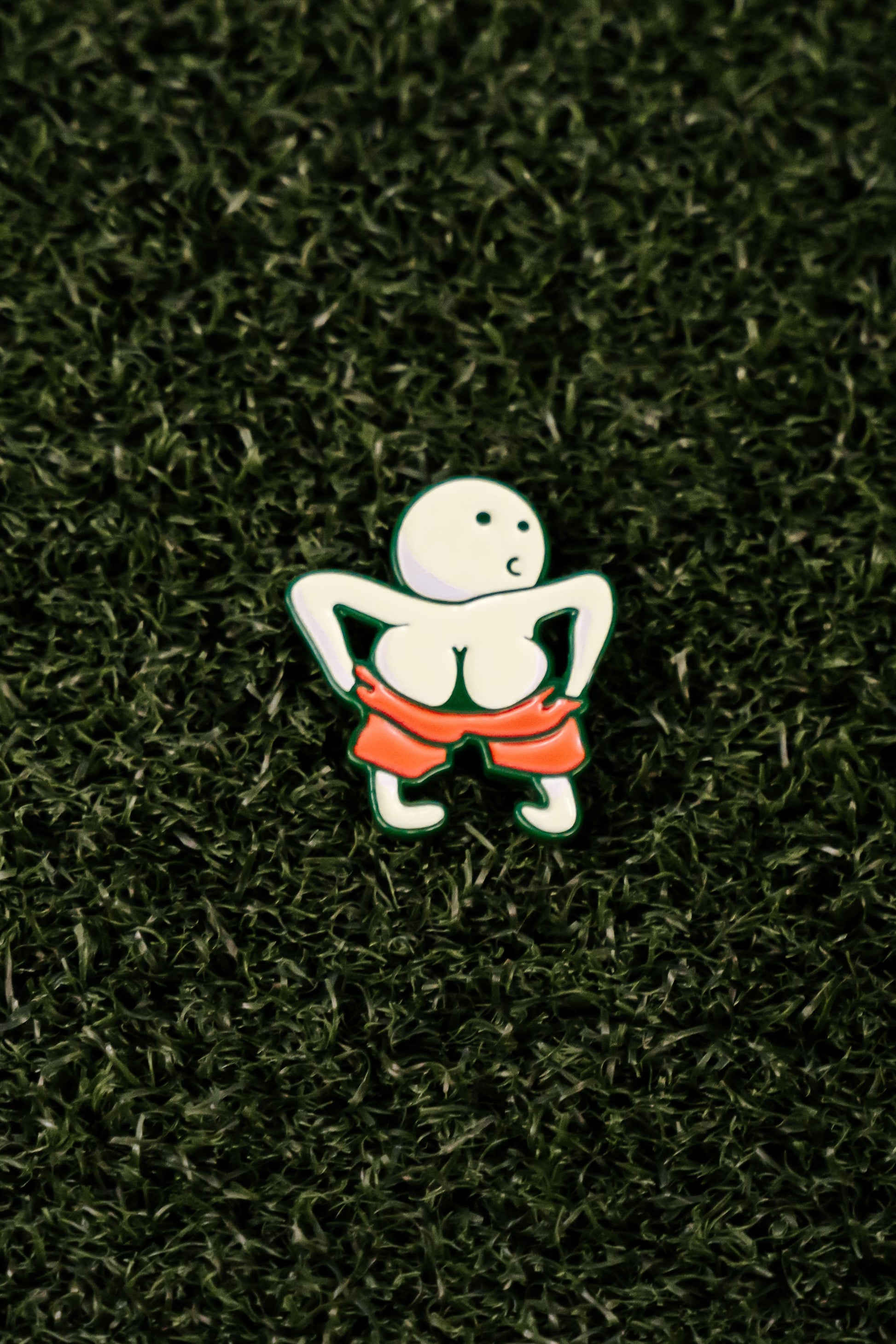The Moon Man Ball Marker by Shank it Golf is a premium metal pin featuring a round-headed figure pulling down red shorts, shown on a dark green turf-like background—ideal for collectors or as a unique magnetic golf accessory.