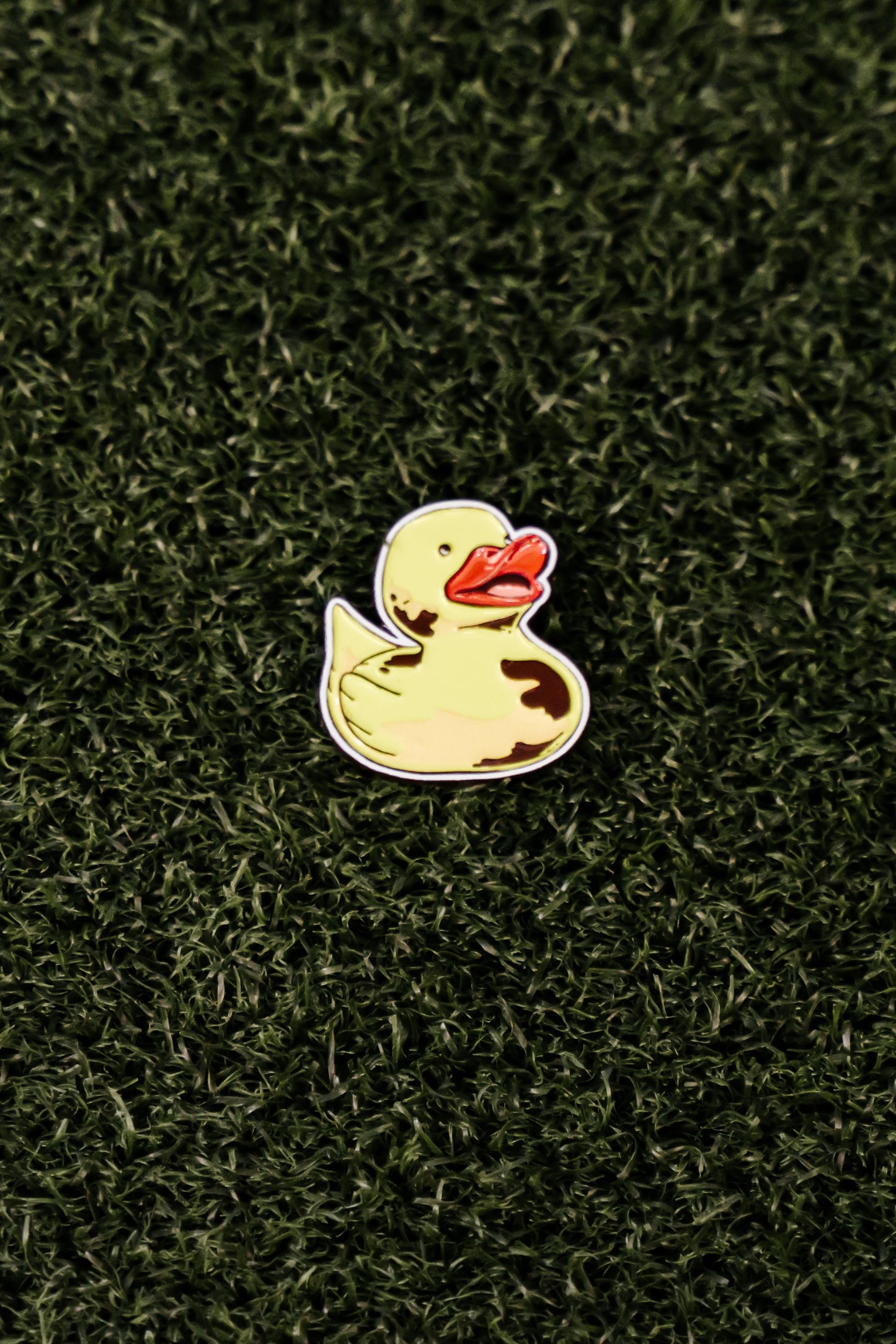 The Ducky Ball Marker by Shank it Golf, featuring a yellow duck with a red beak, is crafted from premium metal and rests on a grass-like surface—perfect as a fun magnetic ball marker for your game.