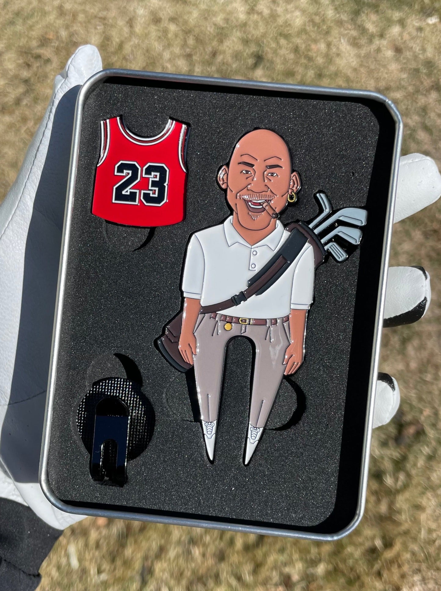 A gloved hand holds "The GOAT 23" pin set by Pin Creatures: a red #23 basketball jersey, a smiling bald man with an earring and golf bag, and a black ball marker, displayed on grass.