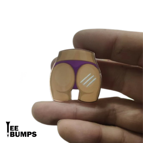 A hand holds the Rack on the Back pin from TeeBumps, shaped like a cartoon lower torso in purple underwear with scratch marks—resembling a banger ball marker. The TeeBumps logo is in the bottom left corner.