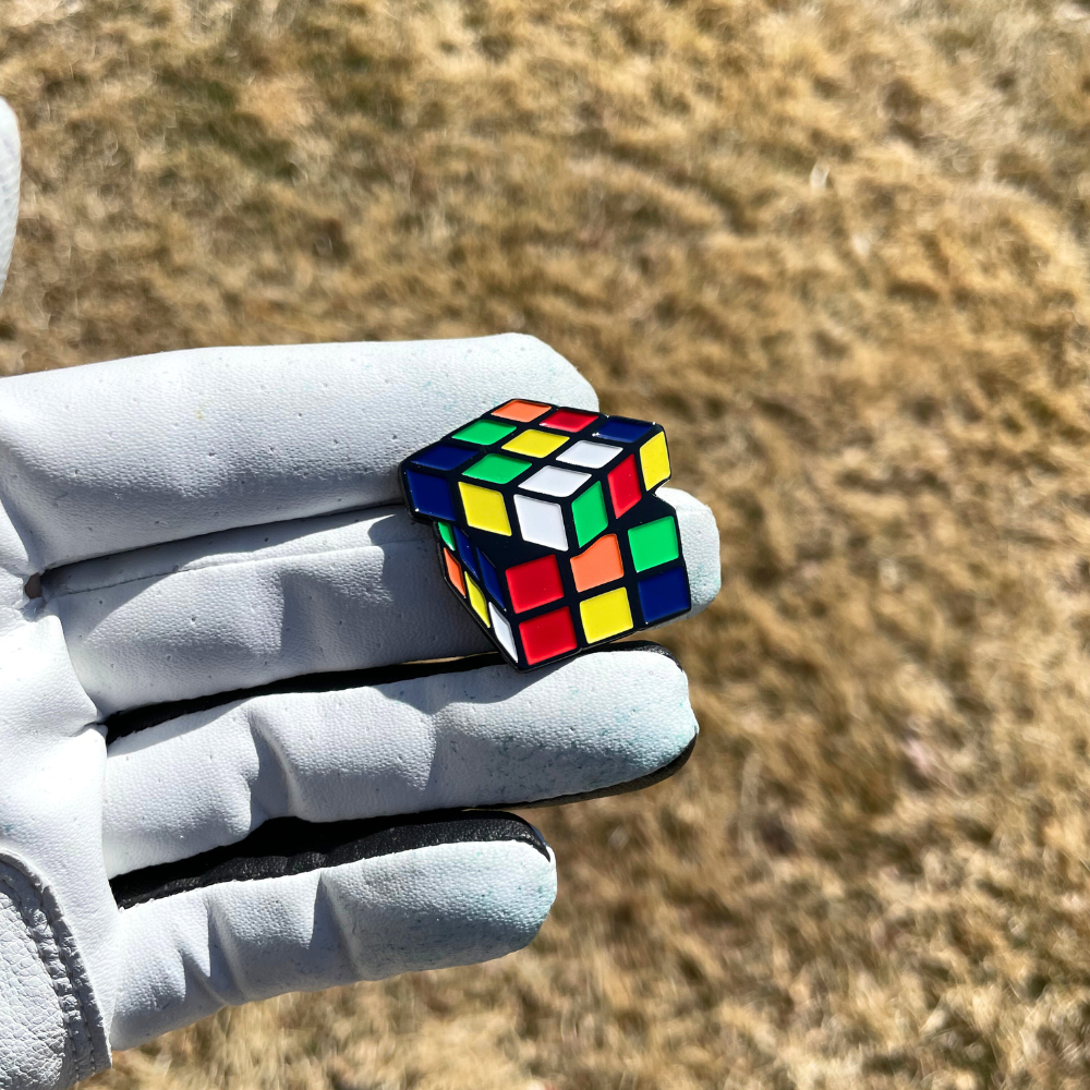 A gloved hand holds a Pin Creatures Rubik's Cube Ball Marker, partly solved, with dry grass softly blurred in the background.