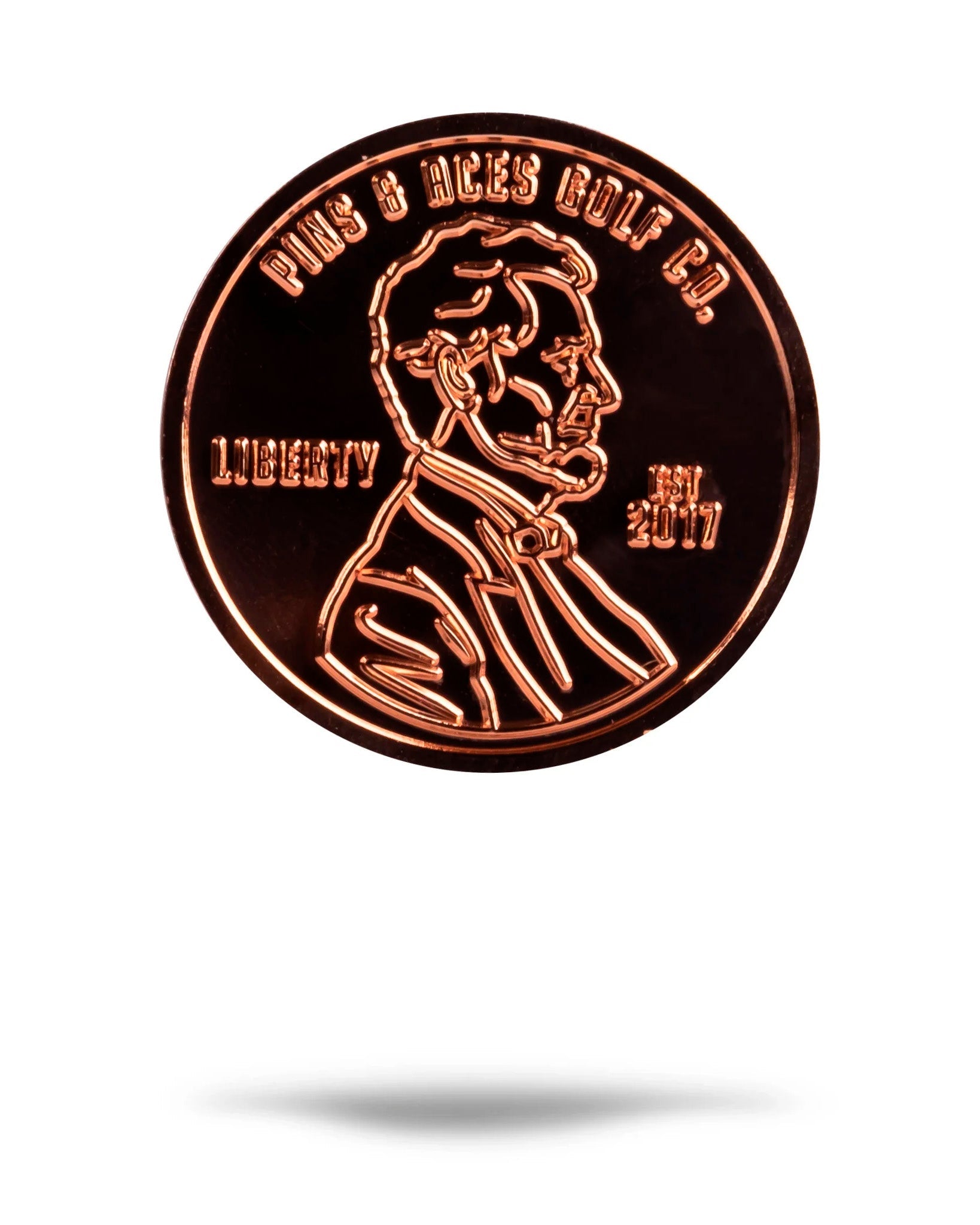 The Pins and Aces Penny Ball Marker is a shiny copper-colored marker with Abraham Lincoln's profile and engraved text: Pins & Aces Golf Co., LIBERTY, and EST 2017. The coin casts a shadow below on a white background.