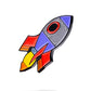 The Rocket Emoji Ball Marker by Pins and Aces is a vibrant enamel pin with red, yellow, purple, and blue accents and a black outline—perfect for golf lovers as both a unique accessory and functional ball marker.