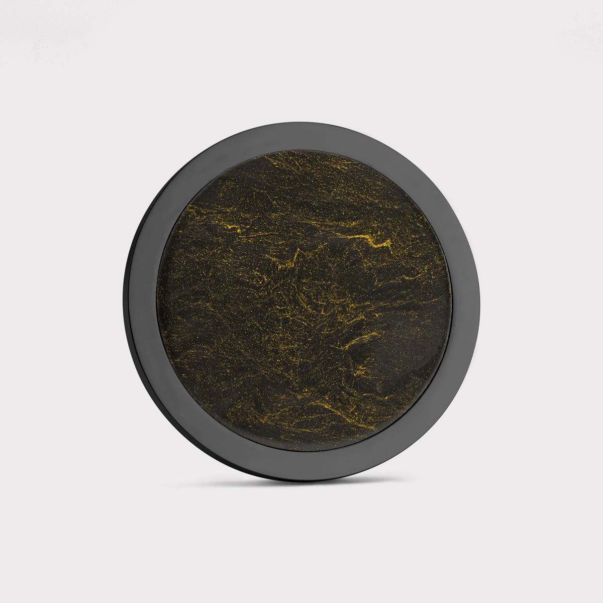 The Venom by Doubloon is a round object with a black outer rim and textured dark surface, highlighted by gold streaks and speckles. Made from forged Carbon Fiber and lightweight Titanium, it’s shown against a plain white background.