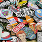 The Custom Mystery Ball Marker Pack features a colorful mix of enamel pins, including food-themed golf ball markers, motorcycles, skulls, faces, mushrooms, and funny signs—all displayed on green grass.