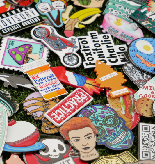 The Custom Mystery Ball Marker Pack features a colorful mix of enamel pins, including food-themed golf ball markers, motorcycles, skulls, faces, mushrooms, and funny signs—all displayed on green grass.