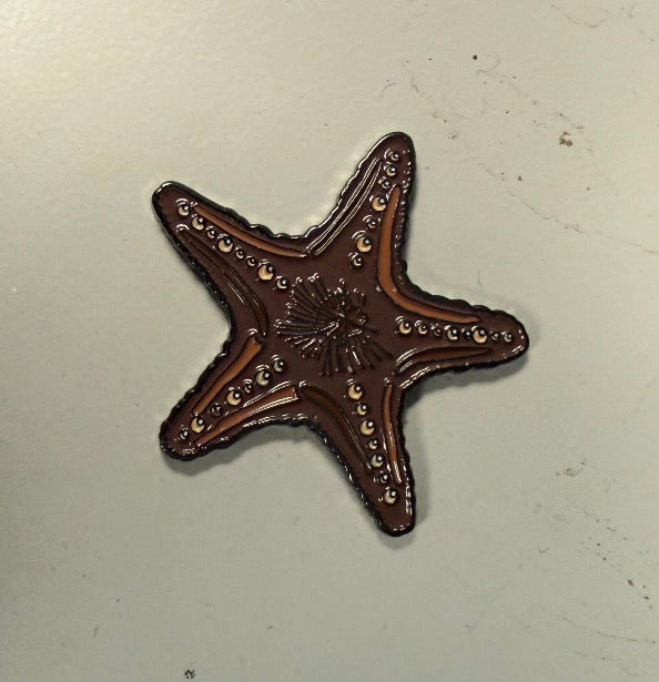 A brown, starfish-shaped Chocolate Starfish Ball Marker from Ballmarkers.com, featuring decorative surface patterns, rests on a plain light-colored background—making it an ideal beach-themed golf accessory for any coastal golf collection.