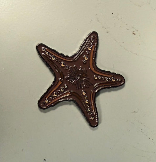 A brown, starfish-shaped Chocolate Starfish Ball Marker from Ballmarkers.com, featuring decorative surface patterns, rests on a plain light-colored background—making it an ideal beach-themed golf accessory for any coastal golf collection.