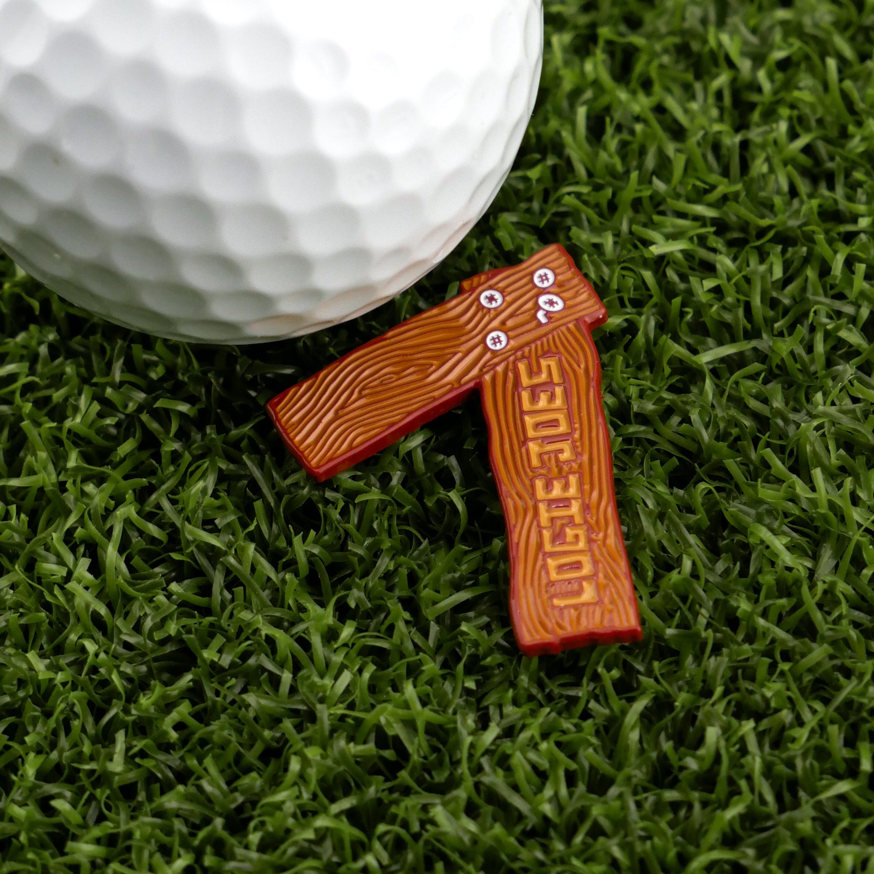 A white golf ball rests on green artificial grass beside Logie Joe's Golf 7 Wood, a small brown axe-shaped premium ball marker with wood grain texture and unique markings—ideal for fans of standout golf accessories.