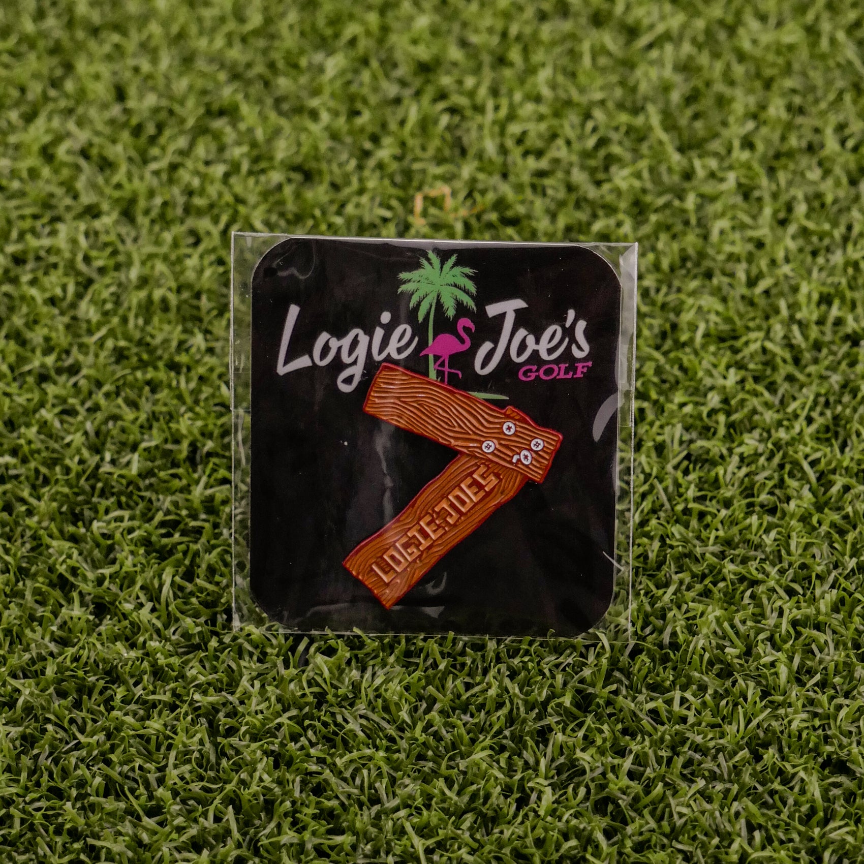 A pin featuring wooden golf clubs and “7 Wood,” by Logie Joe’s Golf, is packaged on a branded card with a palm tree and flamingo, displayed on artificial grass—a unique accessory that doubles as a premium ball marker.