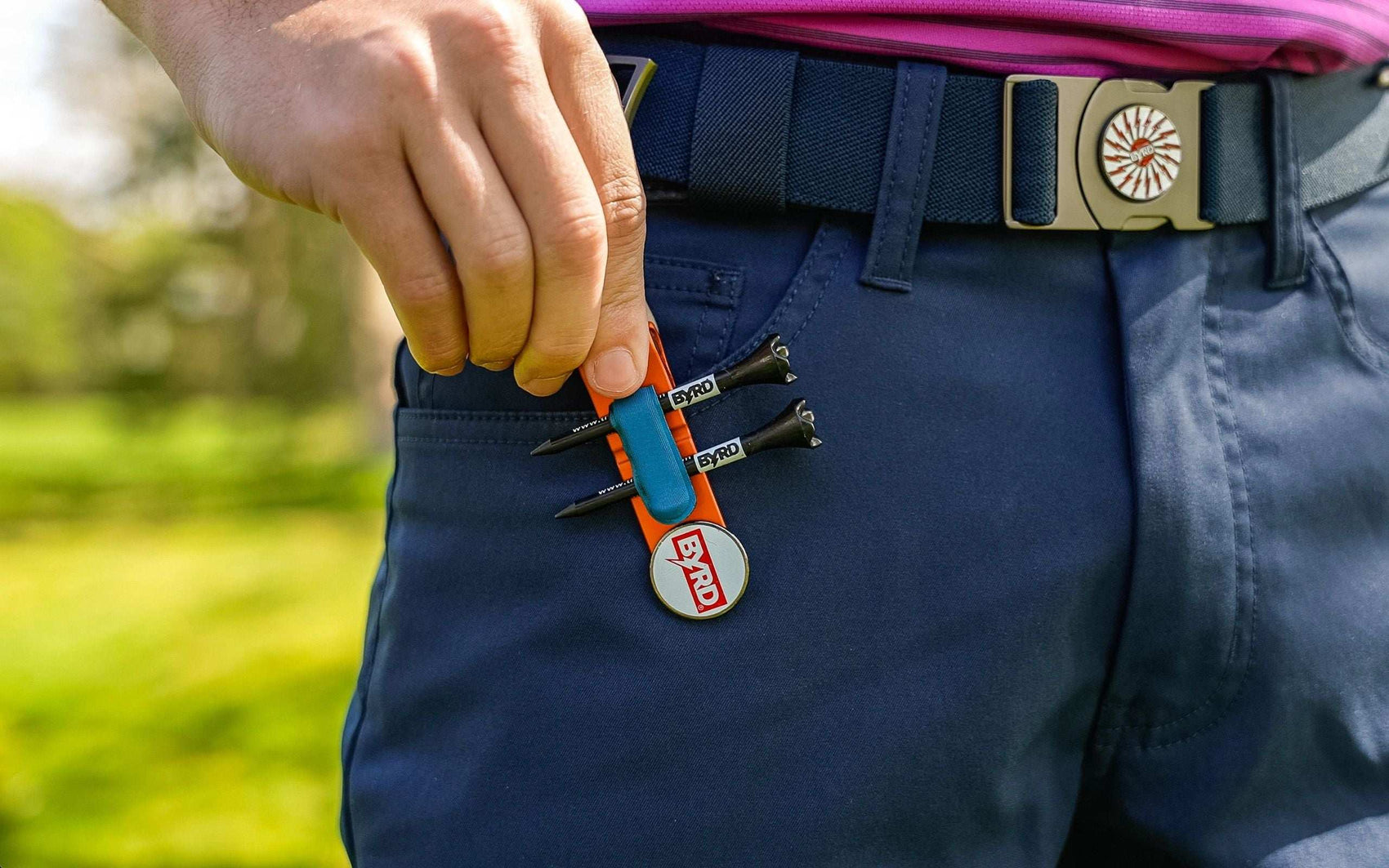 A person in navy blue pants clips The Byrd, LLC’s THE BYRD GOLF CLYP - Orange Oriole with three tees and a BYRD ball marker onto their pocket; a blue belt and pink shirt are also visible.