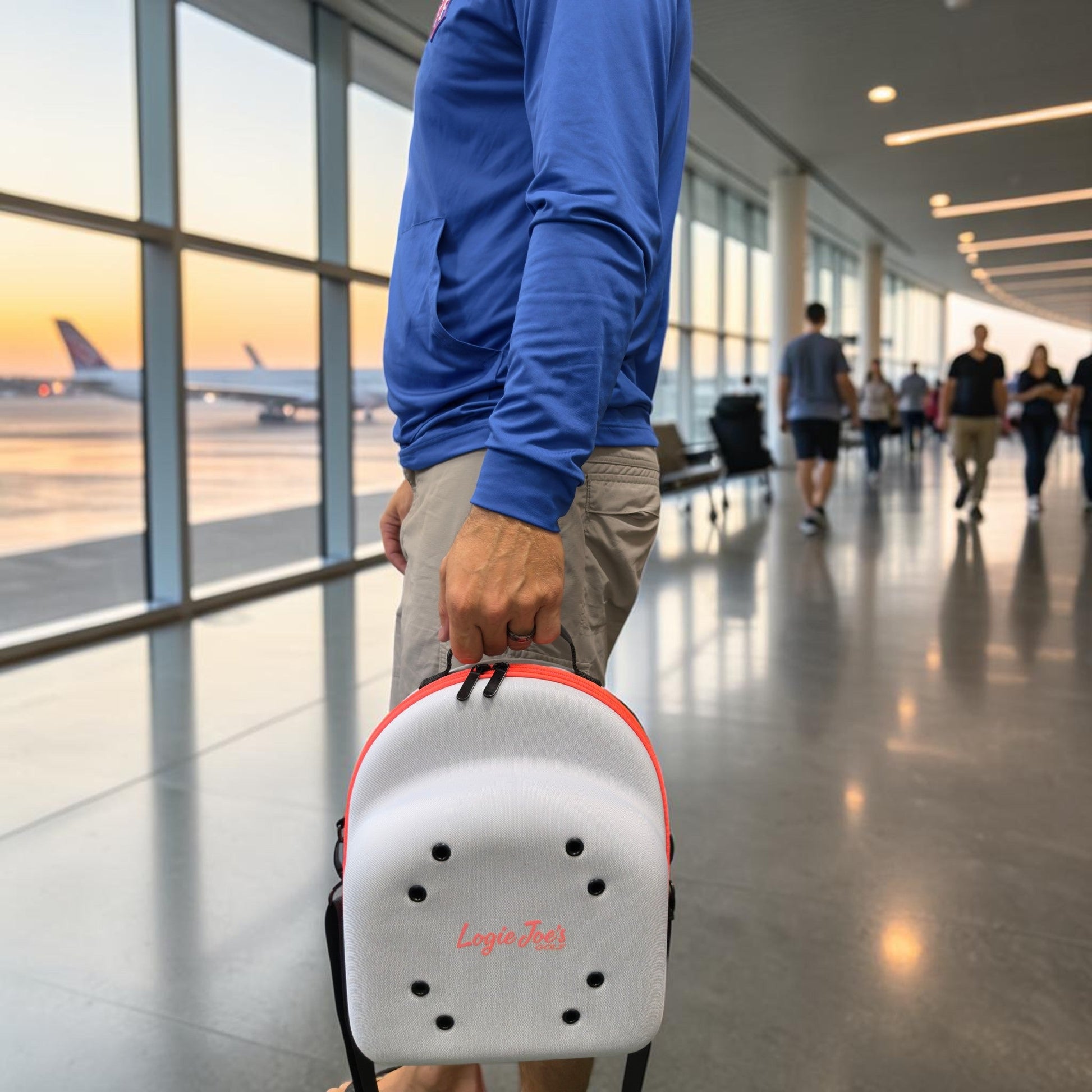 A person in a blue hoodie and khaki shorts stands in an airport terminal near large windows with planes outside at sunset, holding a small white backpack labeled “Logic Jet.” Beside them is the Logie Joe’s Golf Hat Caddy Travel Case—a sleek hard shell hat carrier that holds up to 6 hats, providing protective hat storage and featuring an adjustable shoulder strap for convenient travel.