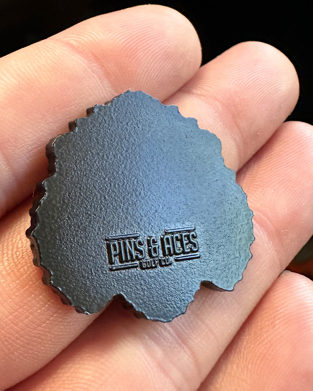 A hand holds the Gopher 2.0 Ball Marker by Pins and Aces, a black, irregularly shaped cast stainless steel item with the embossed logo near the bottom.
