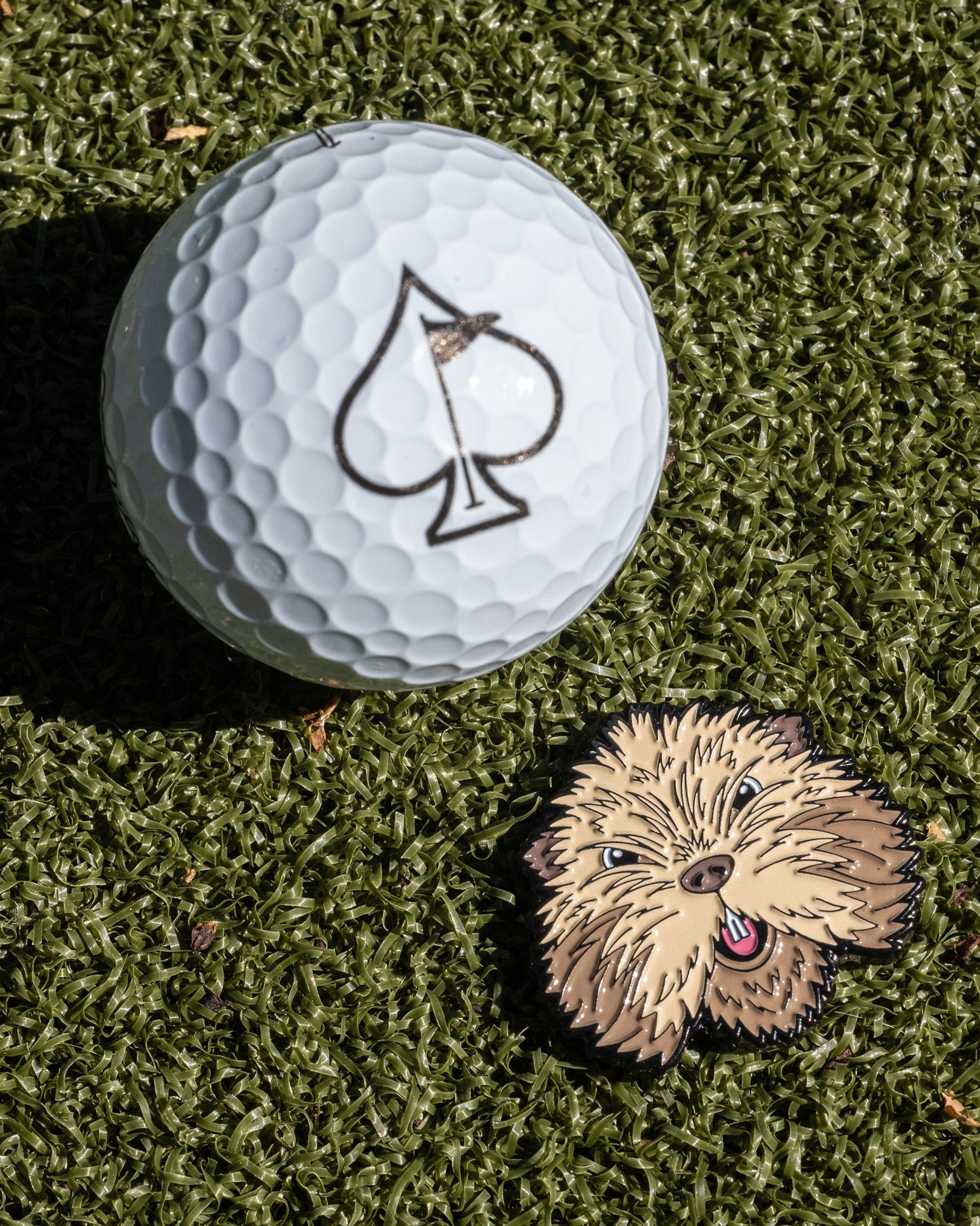 The Pins and Aces Gopher 2.0 Ball Marker features a golf ball with a spade symbol alongside a cute, fluffy brown dog—ideal for fun-loving golfers seeking playful style on the course.