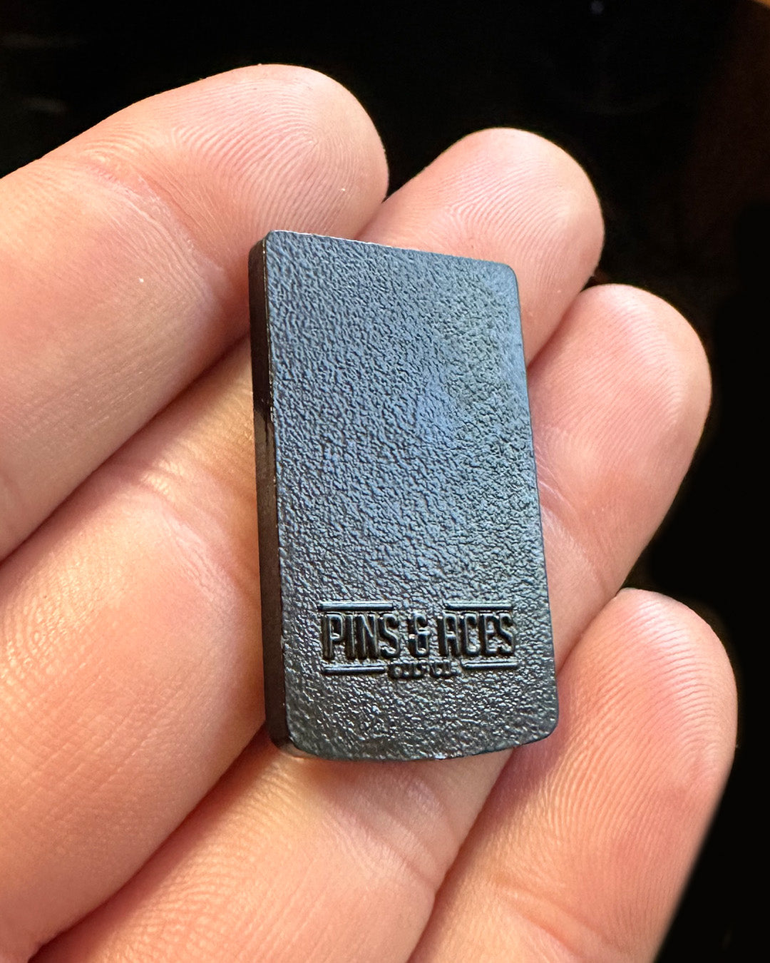 A close-up of a hand holding the Pins and Aces Game Boy Ball Marker—a small, rectangular black accessory with a textured surface and "PINS & ACES" embossed, perfect for golf enthusiasts who appreciate retro gaming style.