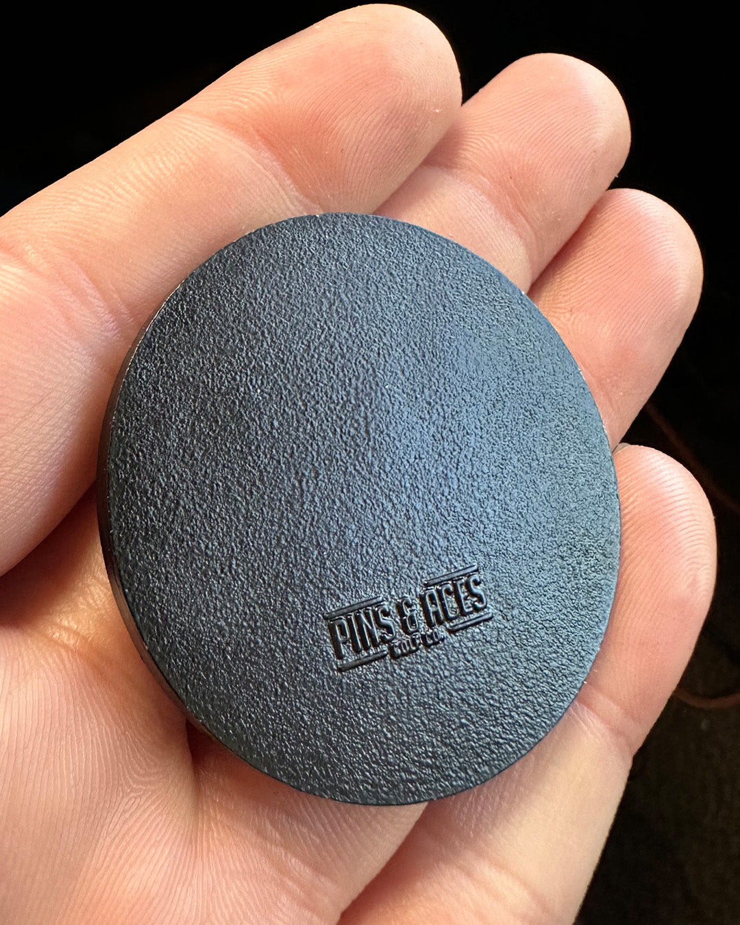 A hand holds a round, flat Golf Ball Ball Marker by Pins and Aces with a textured black surface. Embossed near the bottom edge are PLACES+FACES and STAY YOU, making this magnetic marker both functional and stylish.