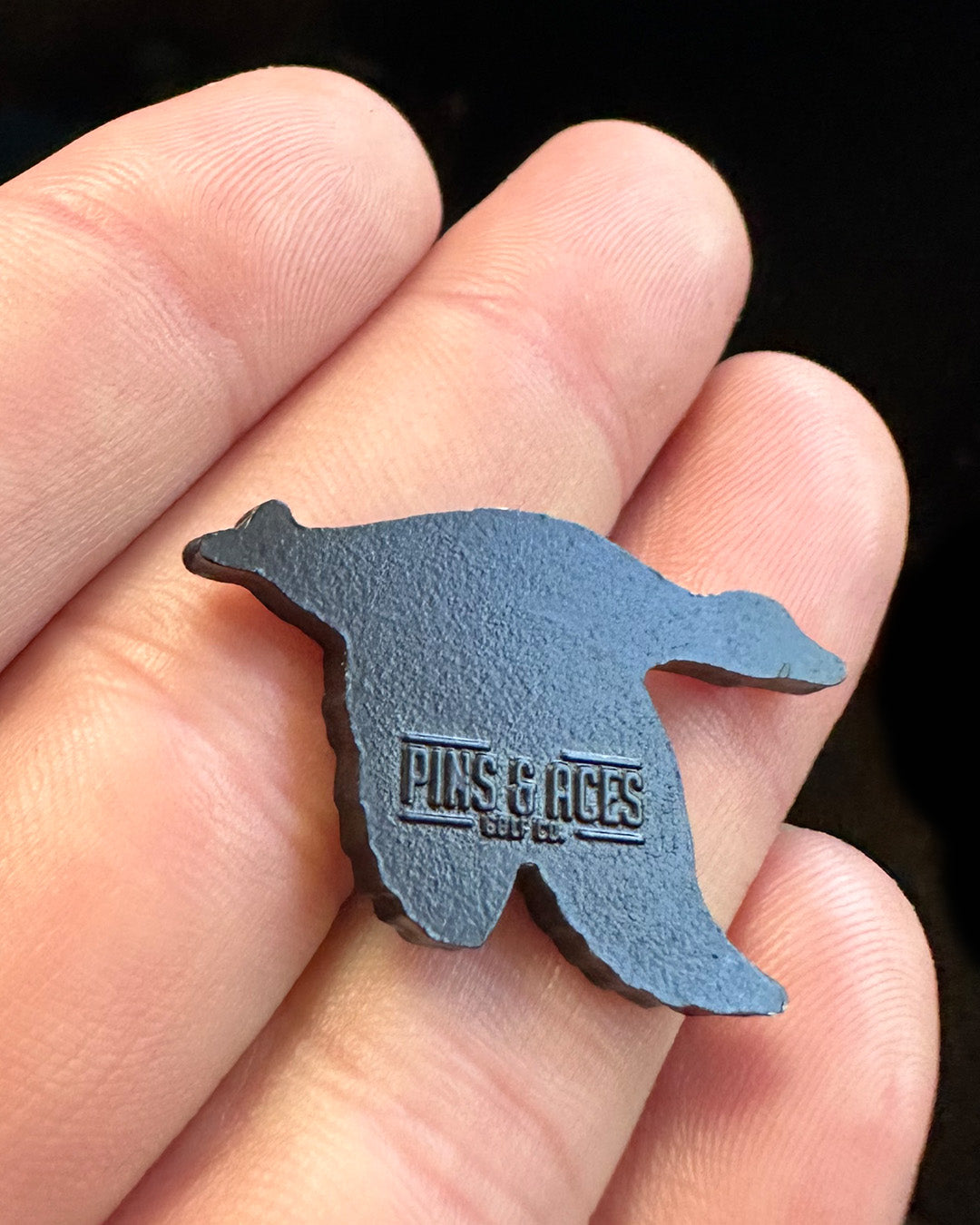 A close-up of a hand holding a Flying Mallard Ball Marker by Pins and Aces, shaped like a mallard and featuring glossy enamel paint with the brand name engraved on its back.