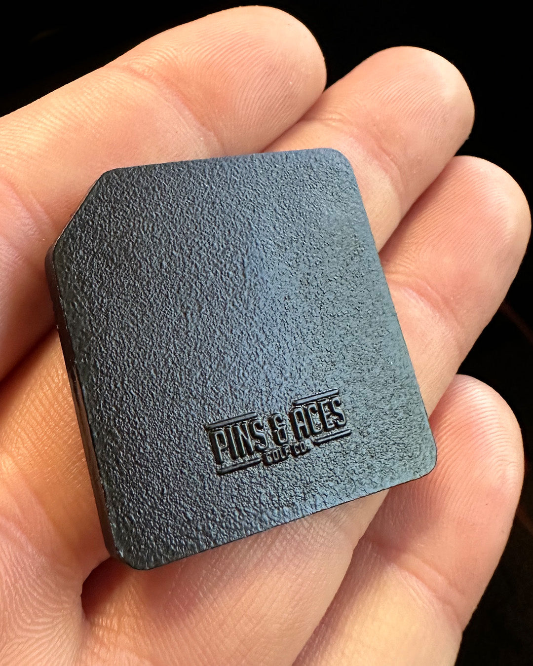 A hand holds the Floppy Disc Ball Marker by Pins and Aces, a small, black, textured square with rounded edges and an embossed Pins and Aces logo—perfect as a golf ball marker for your game.