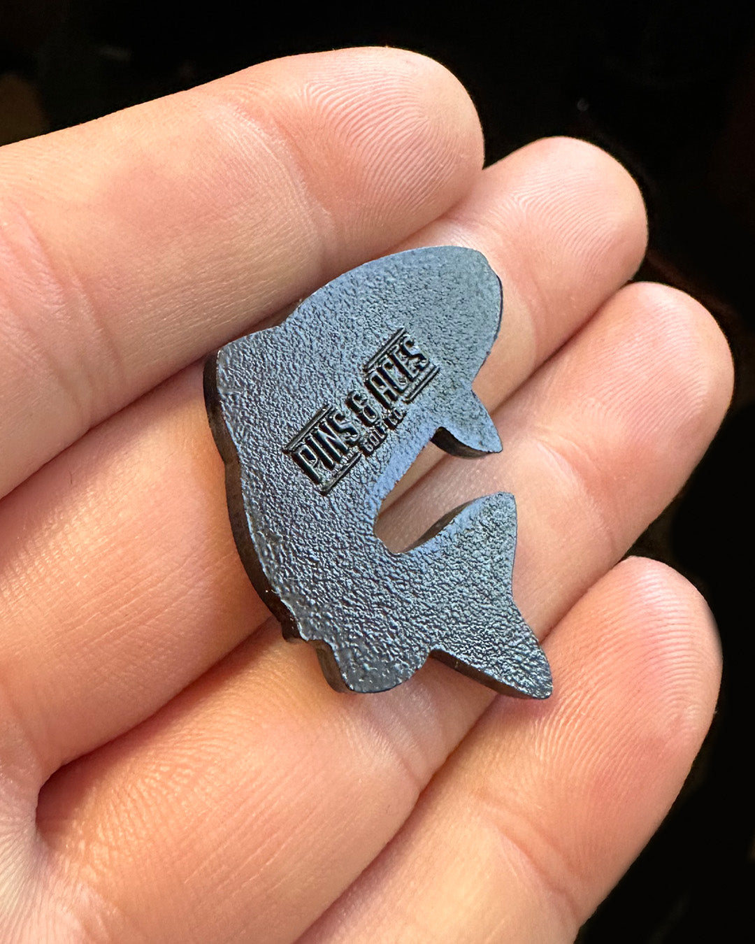 A person holds a small, black Trout Ball Marker by Pins and Aces, shaped like a fish with the brand name embossed on its side. The marker rests in their palm, with fingers visible in the background.