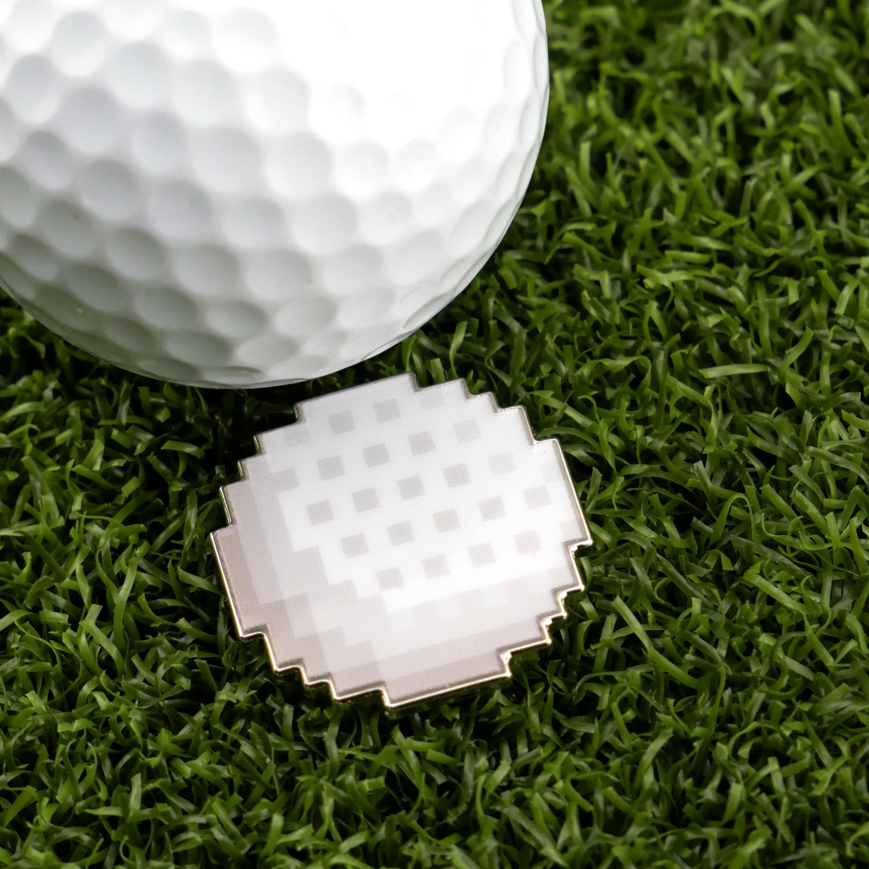 A white golf ball sits on green artificial grass next to the Logie Joe's Golf 8-Bit Golf Ball, a unique, round, jagged-edge accessory that functions as a creative golf ball marker.