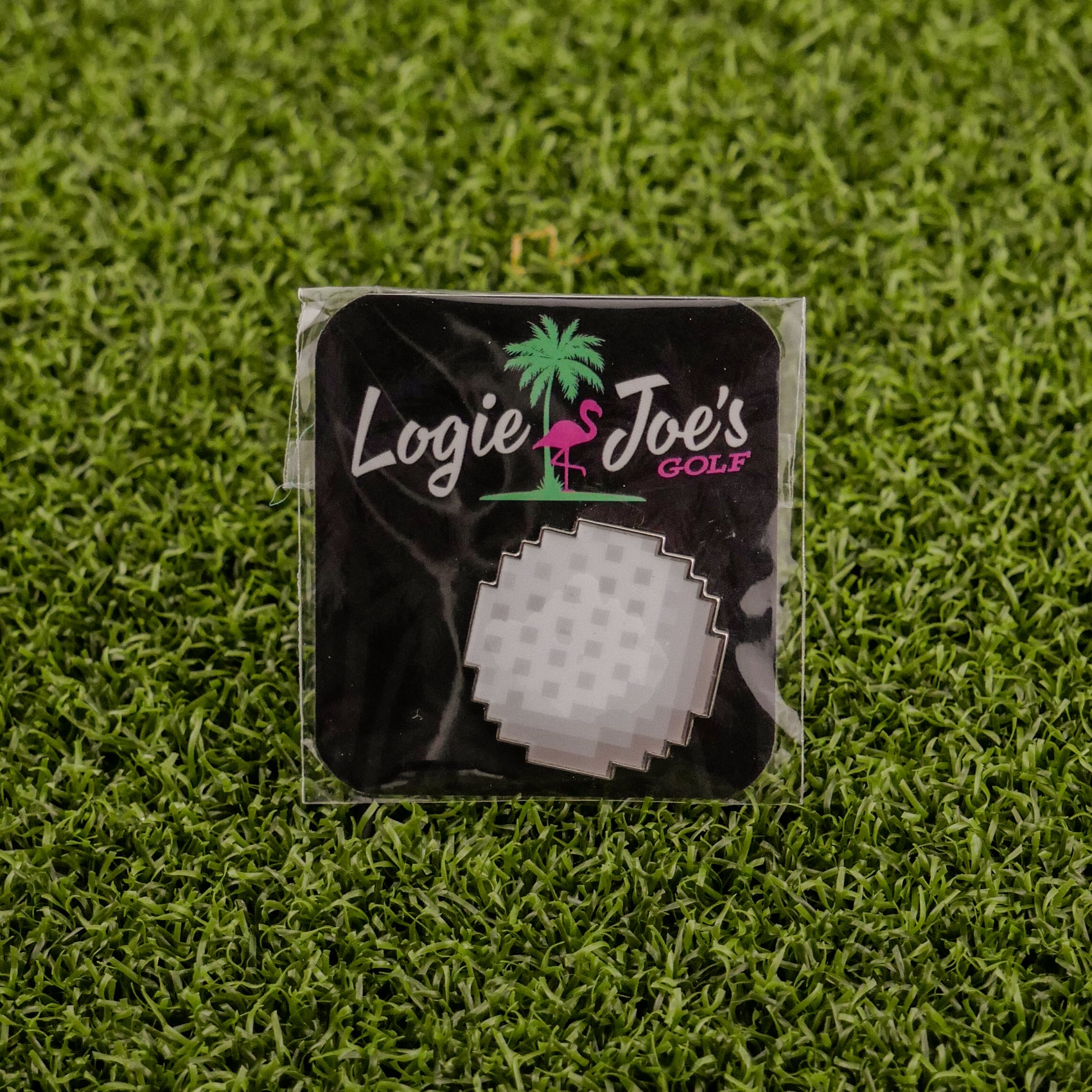 The Logie Joe’s Golf 8-Bit Golf Ball—a pixelated, round white golf ball—appears on green artificial grass in its branded packaging.