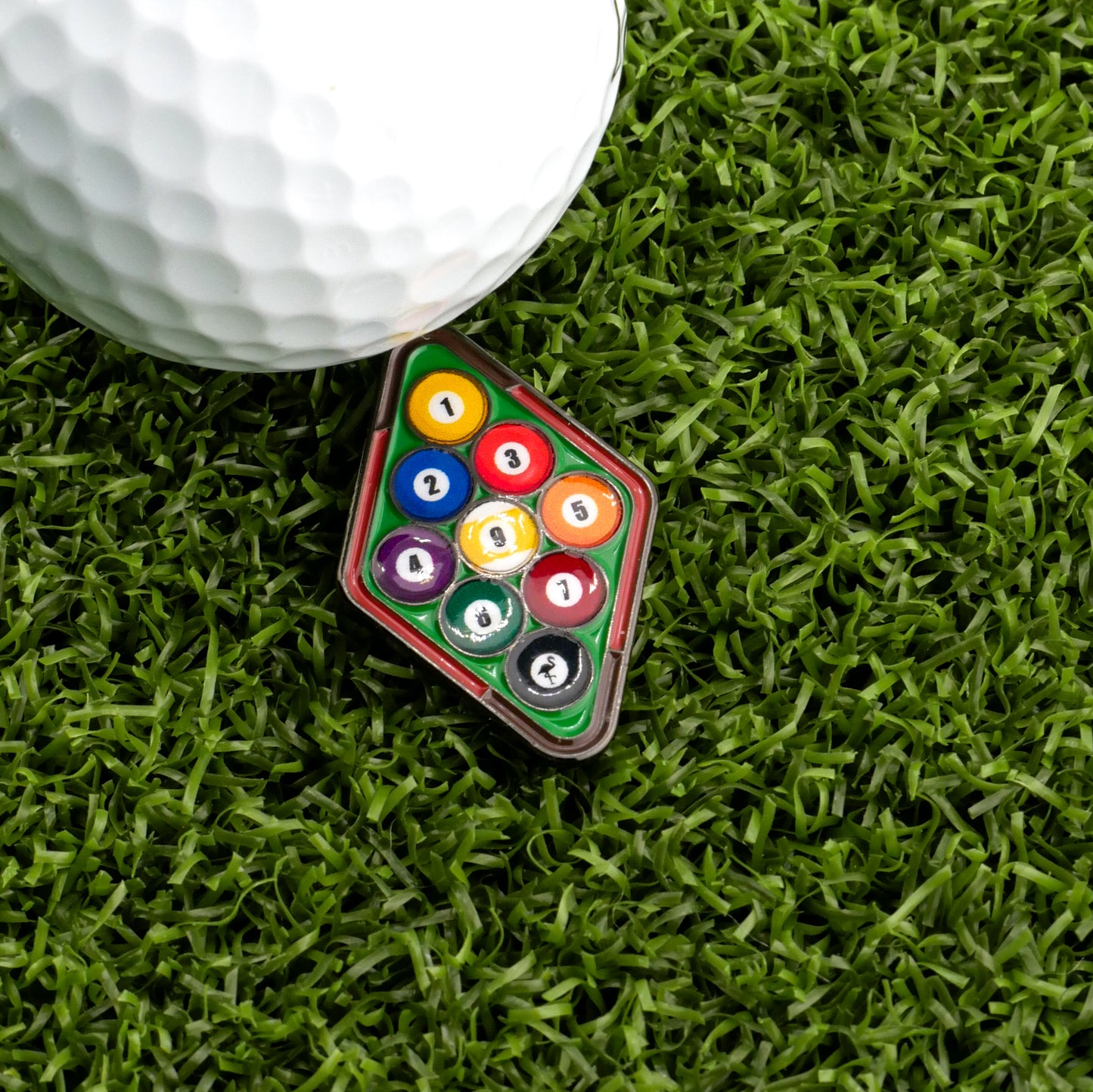 A close-up of Logie Joe's Golf 9-Ball Pool Rack Ball Marker beside a mini triangle rack of billiard balls on green turf—an amusing gift for golf lovers with a sense of humor.