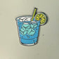 A drawing featured on the Water Hazard Ball Marker by Ballmarkers.com shows a blue drink in a glass filled with ice cubes, topped with a yellow straw and a lime slice on the rim—capturing the refreshing feeling of cooling down after conquering a water hazard on the golf course.