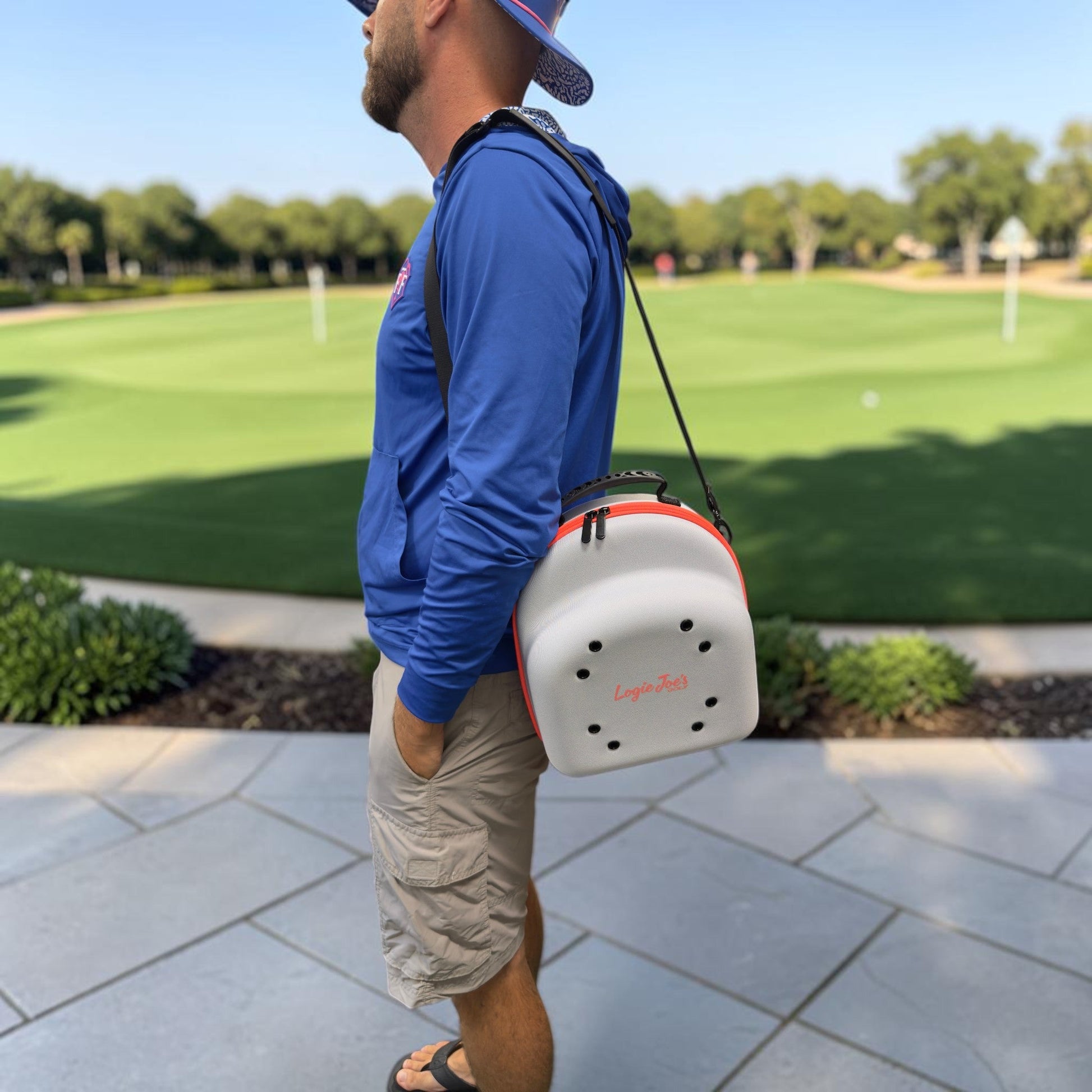 A person in a blue hoodie, tan shorts, sandals, and a hat stands on a paved area by a golf course, carrying a white and red LogoPutt bag over their shoulder along with the Logie Joe’s Golf Hat Caddy Travel Case—a hard shell hat carrier that holds up to 6 hats—for added convenience.