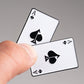 A close-up of a hand holding two aces of spades playing cards, with the Pins and Aces Ace of Spades Ball Marker—a sleek enamel golf accessory—resting nearby against a neutral background.