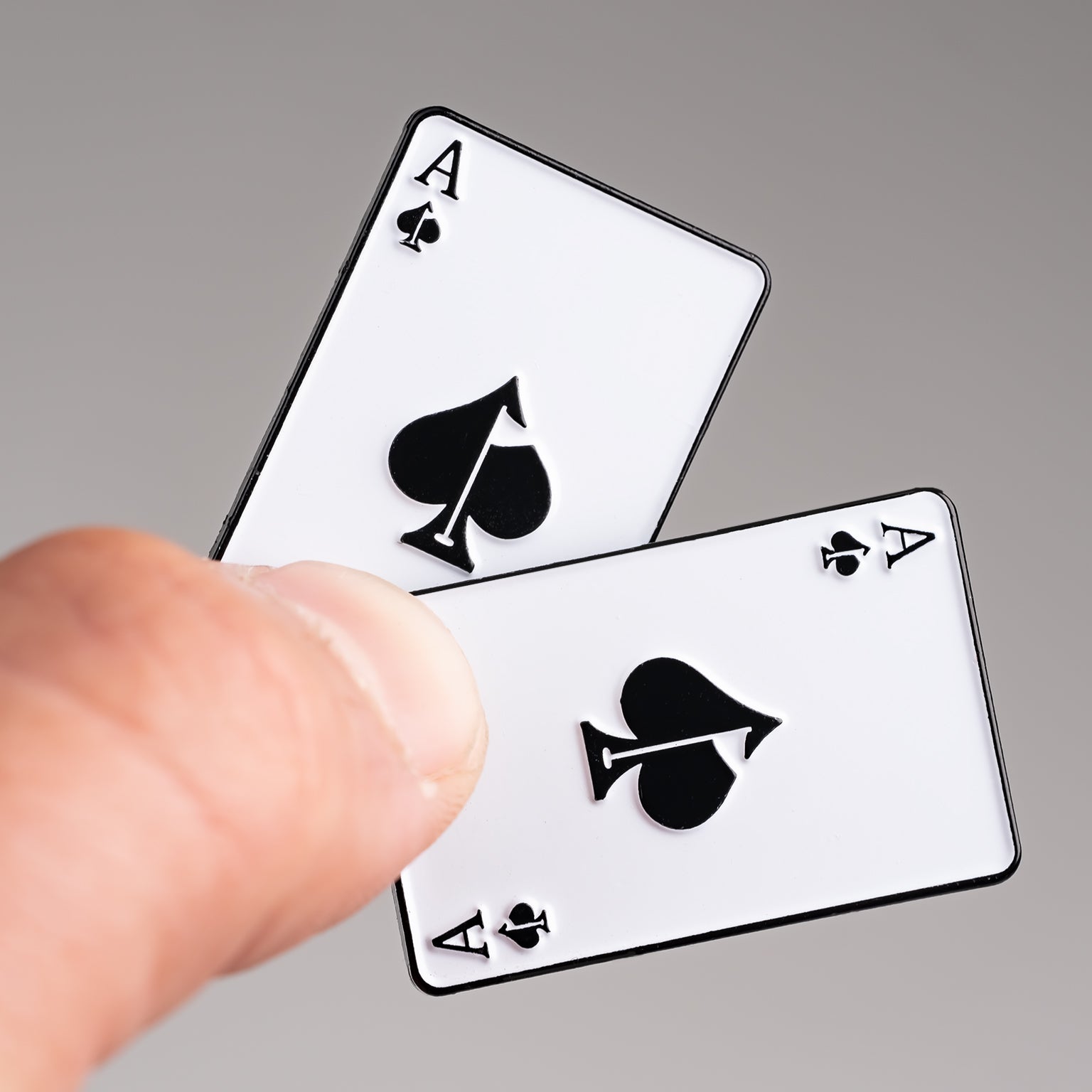 A close-up of a hand holding two aces of spades playing cards, with the Pins and Aces Ace of Spades Ball Marker—a sleek enamel golf accessory—resting nearby against a neutral background.