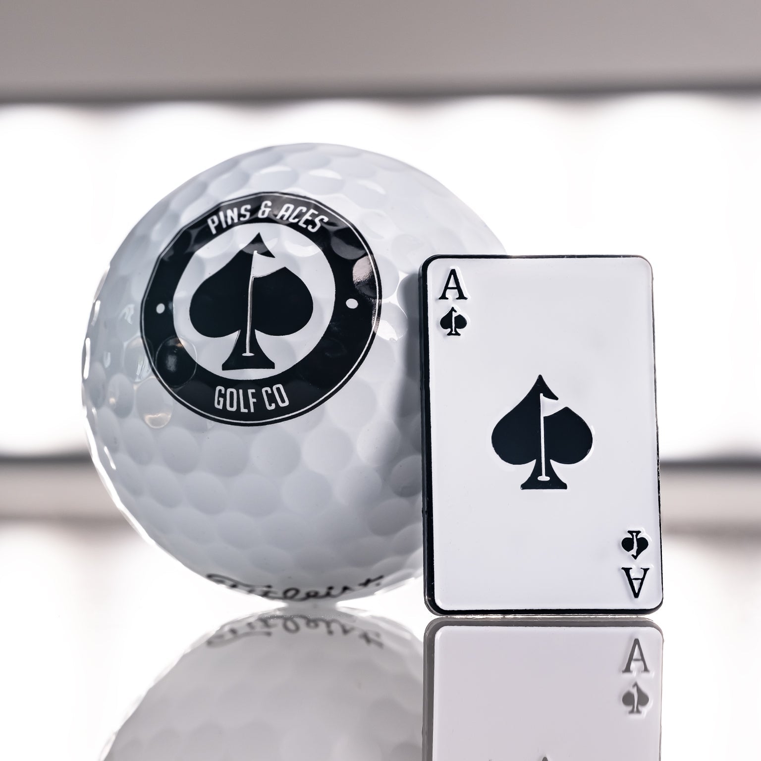 A premium golf accessory: the Pins and Aces Ace of Spades Ball Marker, shaped like the ace of spades, alongside a white golf ball with a black spade logo—both elegantly reflecting on a gleaming surface.