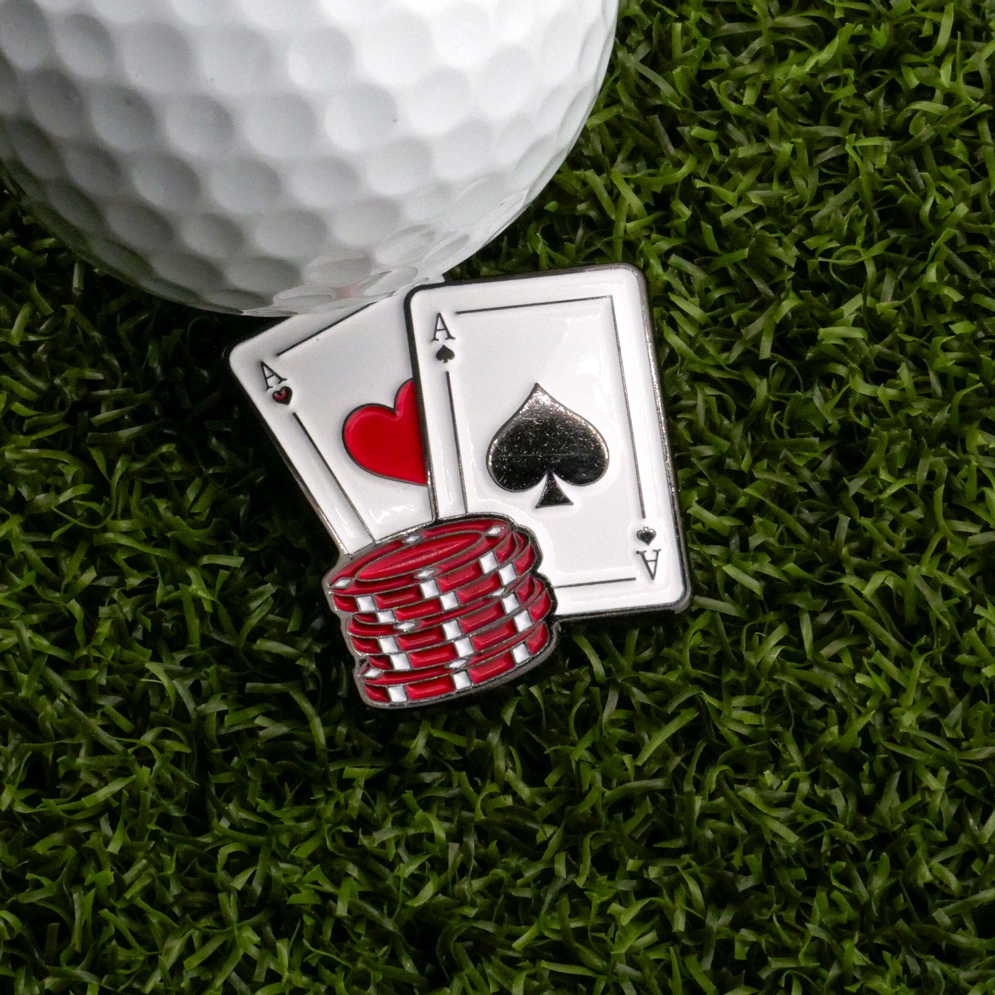 A close-up of Ballmarkers.com's Big Pair of Aces Ball Marker beside a golf ball, ace of hearts, ace of spades, and red poker chips on green grass.