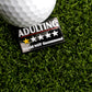 The Ballmarkers.com Adulting Sucks Ball Marker features a golf ball on artificial grass next to a pin that says ADULTING with five stars—just one gold—and "Would NOT Recommend," humorously highlighting golf as an escape from adult life.