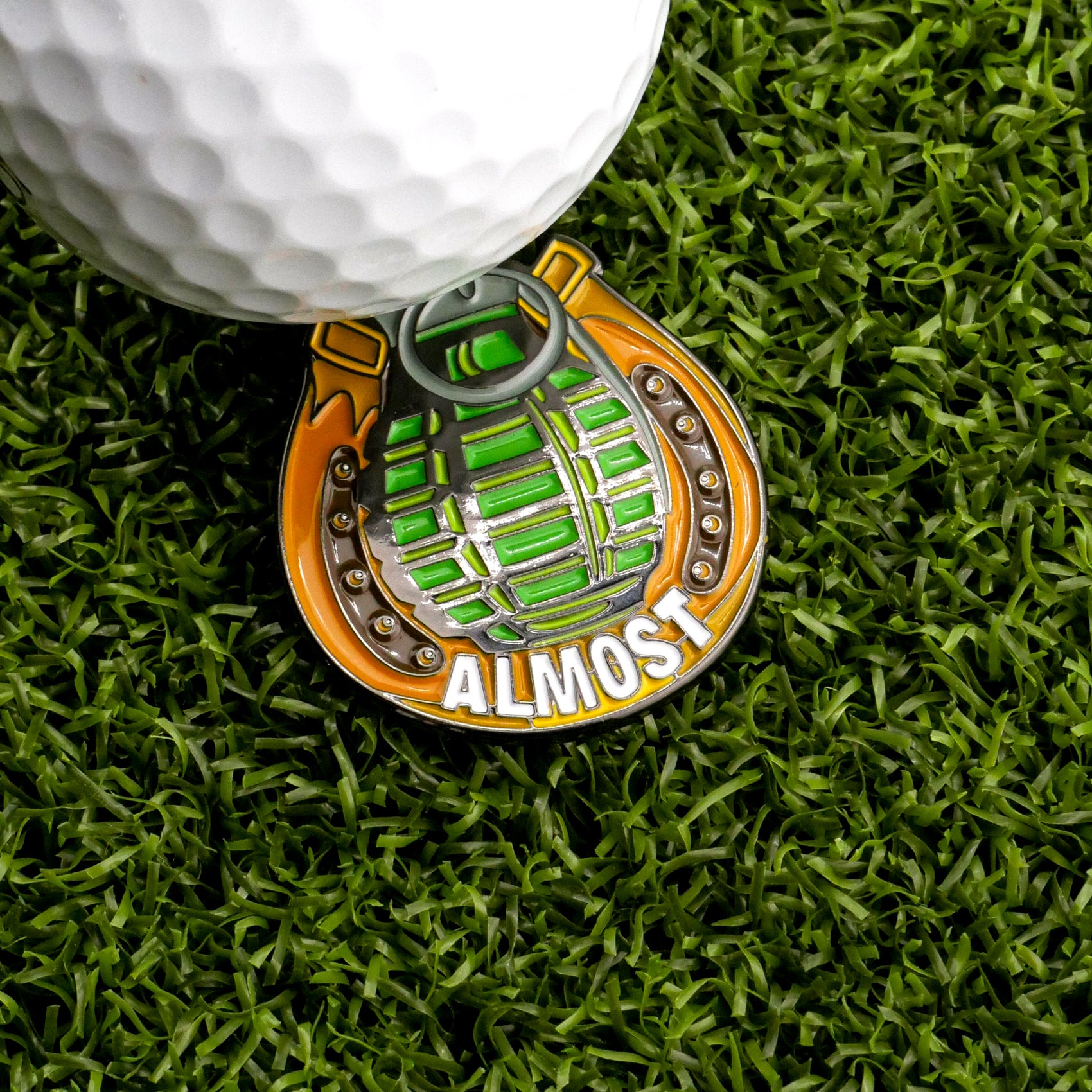 The Almost Only Counts Ball Marker by Ballmarkers.com sits on green artificial grass next to a golf ball—its hand grenade shape and bold "ALMOST" lettering make it a standout accessory for any golfer.