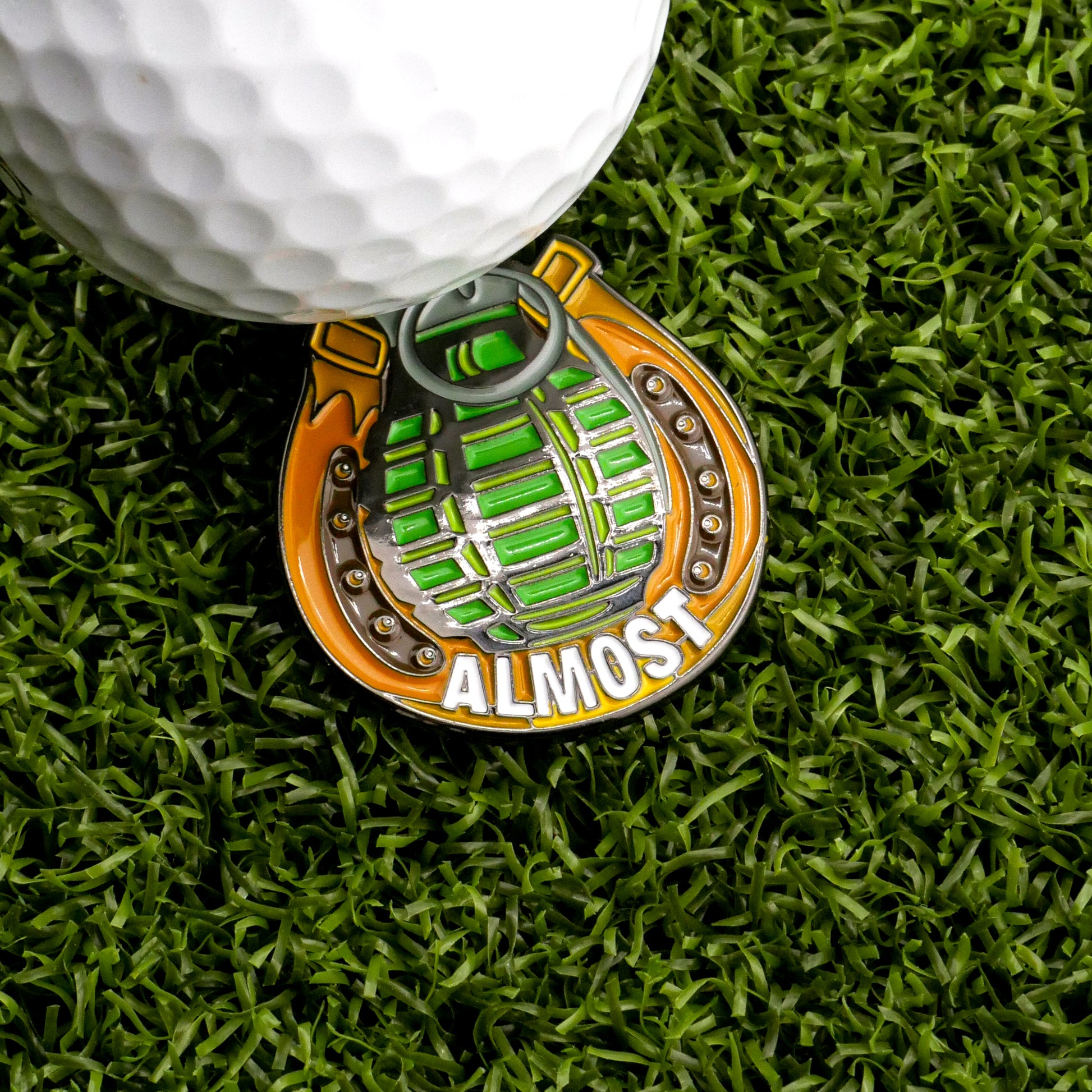 The Almost Only Counts Ball Marker by Ballmarkers.com sits on green artificial grass next to a golf ball—its hand grenade shape and bold "ALMOST" lettering make it a standout accessory for any golfer.