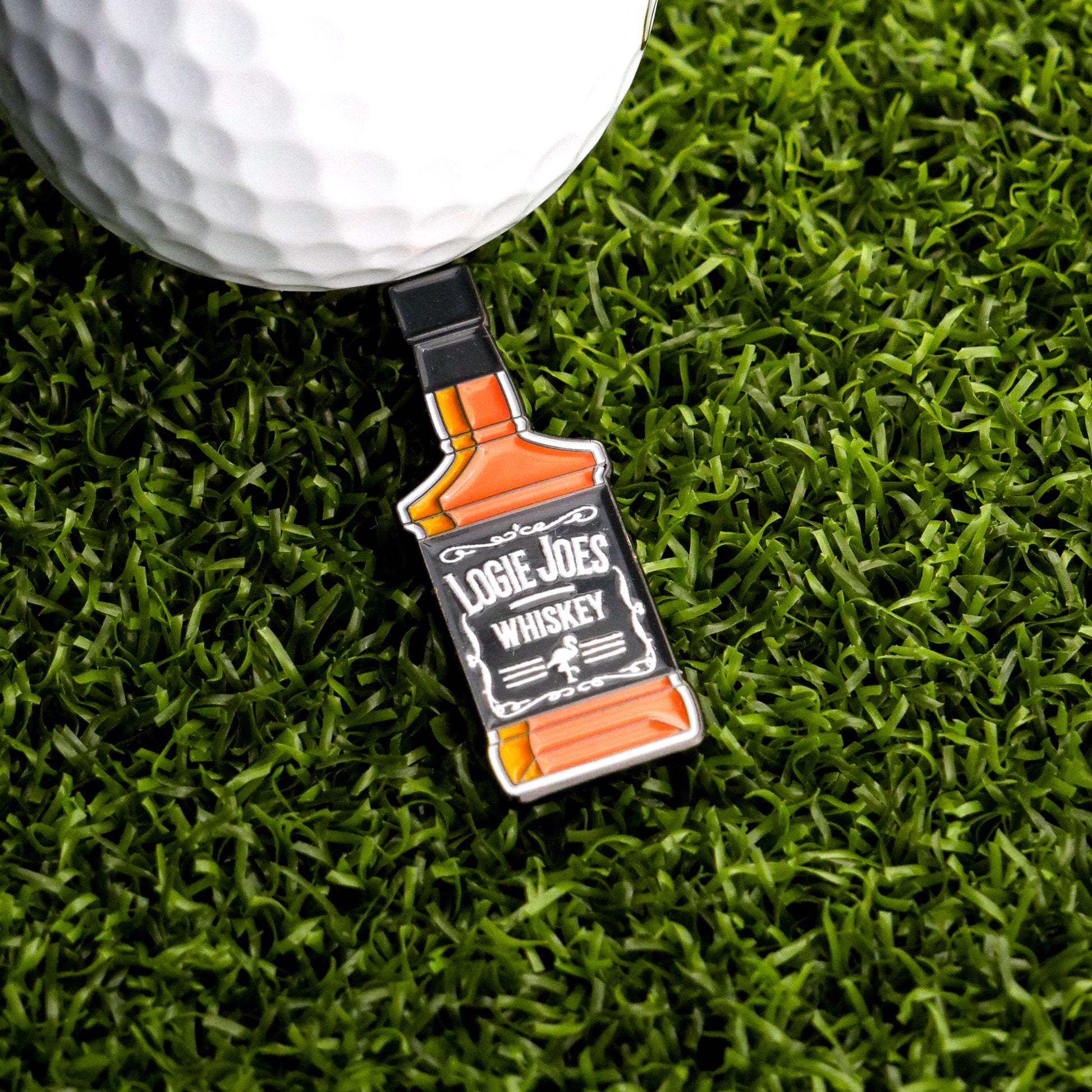 A golfer's ball rests on artificial grass beside Logie Joe's Golf American Whiskey Ball Marker, shaped like a bottle and hinting at an appreciation for whiskey.