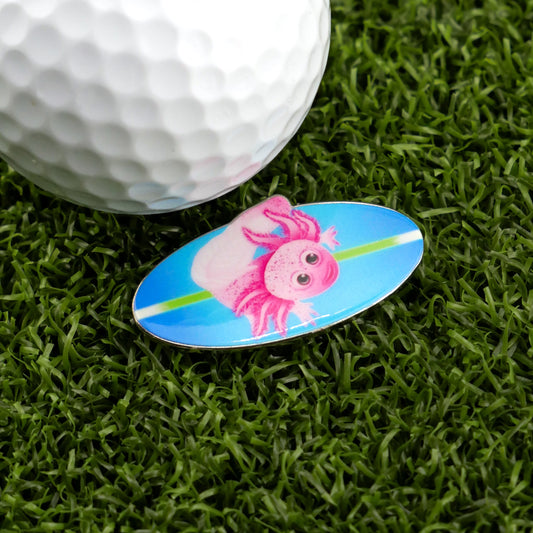 A golf ball on artificial grass sits beside the Logie Joe's Golf Axolotl Ball Marker—an oval with a pink axolotl illustration on a blue background.