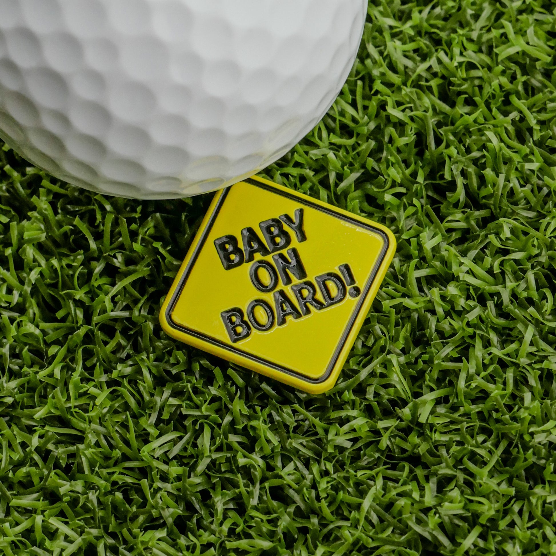 The Logie Joe's Golf Baby On Board Ball Marker, a funny yellow golf accessory, is shown on artificial grass beside a white golf ball.