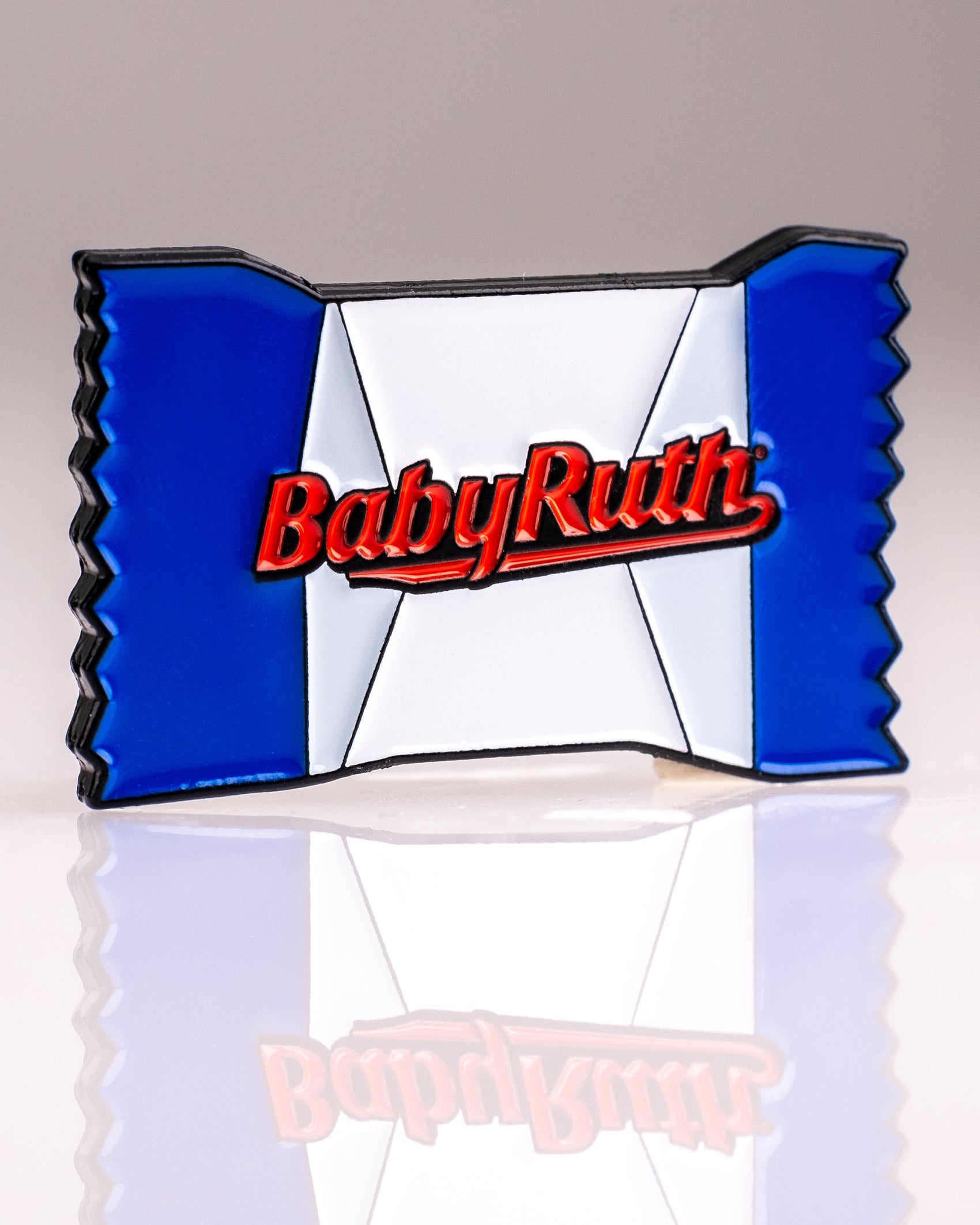 The Baby Ruth® Ball Marker from Pins and Aces is a limited edition enamel pin styled like a Baby Ruth bar in a blue, white, and red wrapper with "Baby Ruth" in red, perfect as both a collectible pin and golf ball marker. Photographed on a reflective surface.