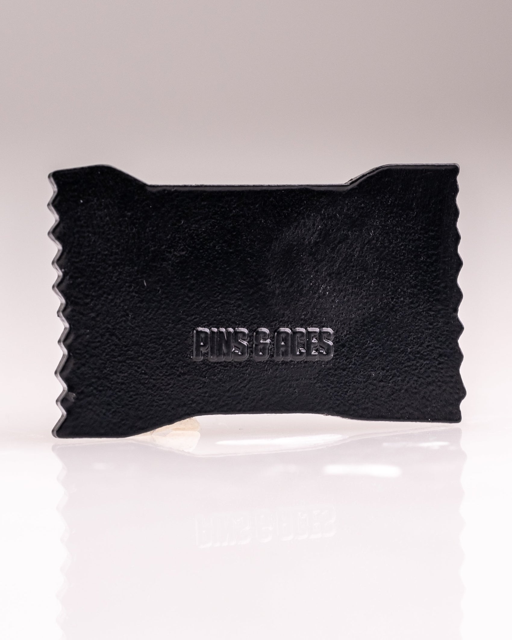 A black, rectangular Baby Ruth® Ball Marker by Pins and Aces features zigzag edges and embossed branding, photographed on a white background. This limited edition accessory brings style to your golf game.