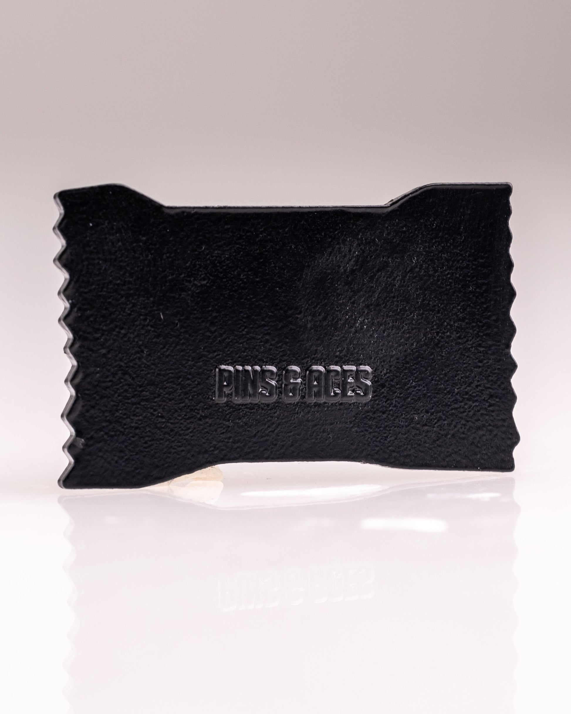 A black, rectangular Baby Ruth® Ball Marker by Pins and Aces features zigzag edges and embossed branding, photographed on a white background. This limited edition accessory brings style to your golf game.