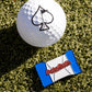 A white golf ball with a black spade sits on green turf next to the limited edition Pins and Aces Baby Ruth® Ball Marker, designed to resemble a blue and white candy wrapper.