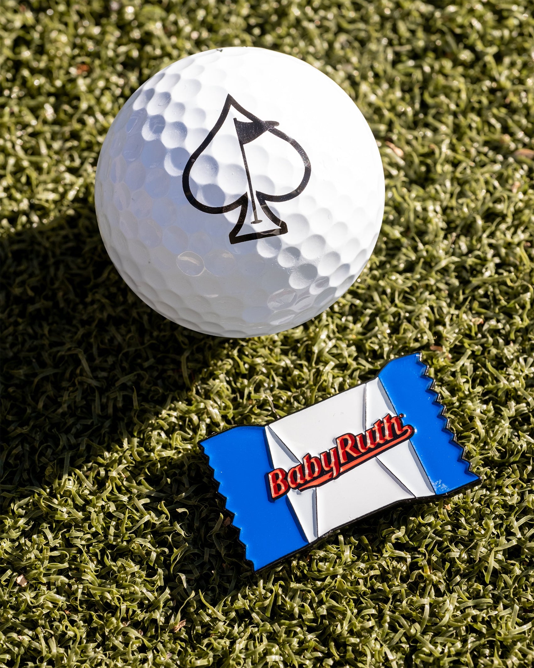 A white golf ball with a black spade sits on green turf next to the limited edition Pins and Aces Baby Ruth® Ball Marker, designed to resemble a blue and white candy wrapper.
