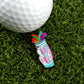 A golf ball sits on green turf beside Logie Joe's Golf “Bag-O-Dicks” marker, shaped like a golf bag and decorated with colorful clubs—a bold, fun accessory for your premium golf collection.
